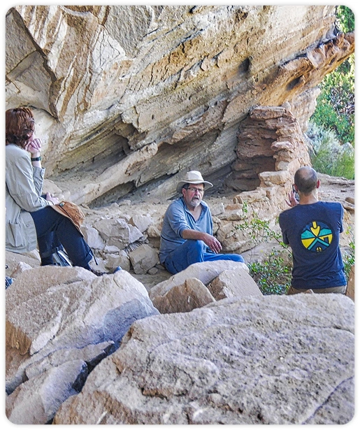 A photograph of several people sitting in the shade of aan alcove near some archeaological ruins.